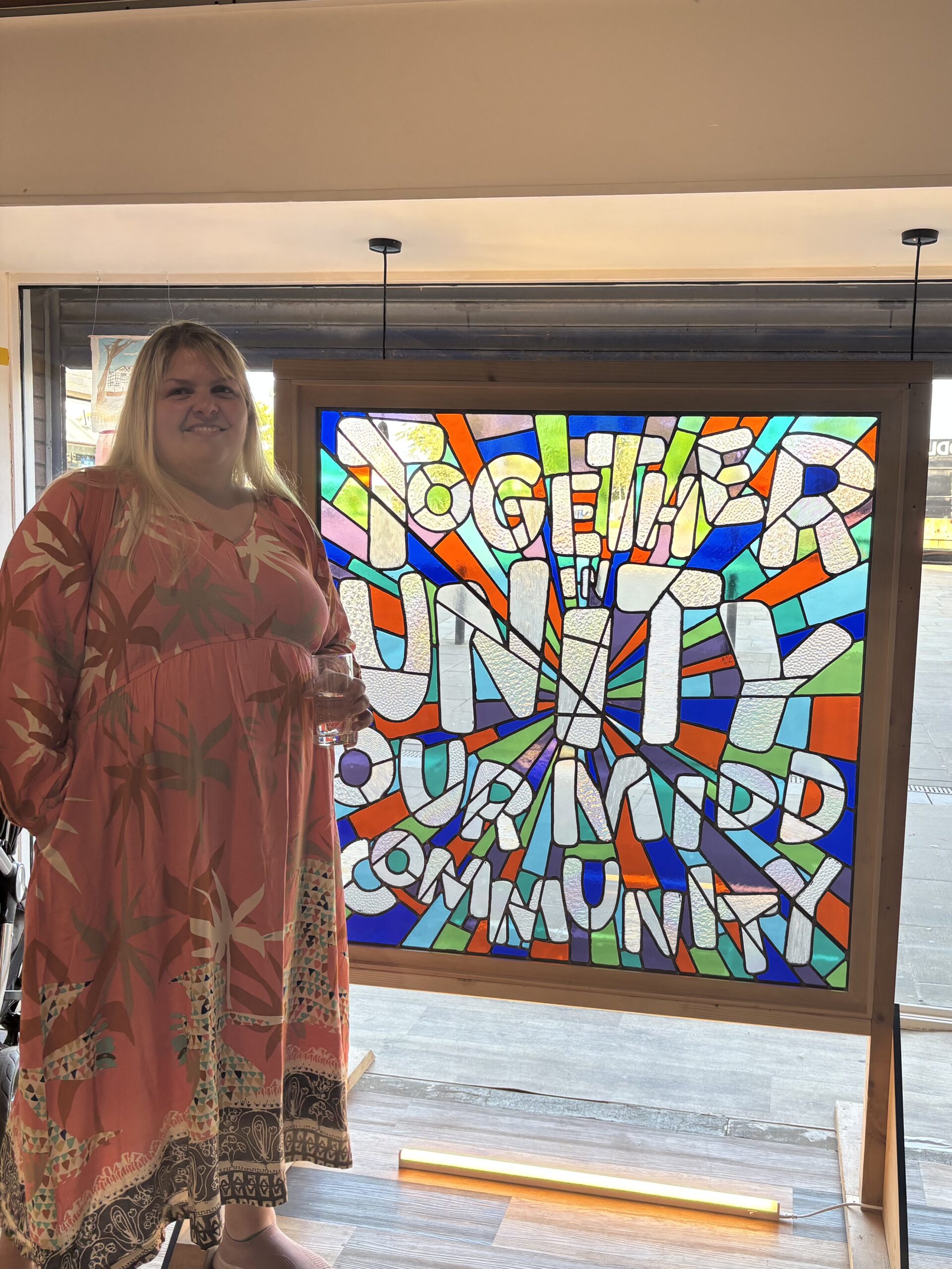 Laura Orton stood next to a stained glass window which reads 'together in unity, our midd community' in multicoloured glass panels